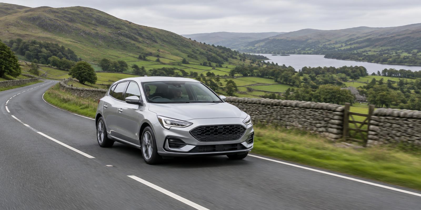 Silver hatchback driving along a country road in the Lake District with stone walls and a lake behind